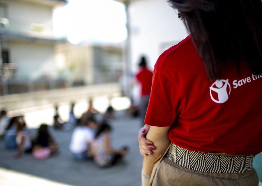 Operatrice save the children con la maglietta ripresa di spalle fino a sotto la vita, guarda bambini seduti a terra ma sfocati nella foto