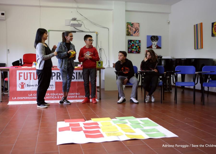 Alcuni bambini e bambine del progetto Save the Children Futuro Prossimo sono in piedi, altri seduti, di fronte a un cartellone con vari post it