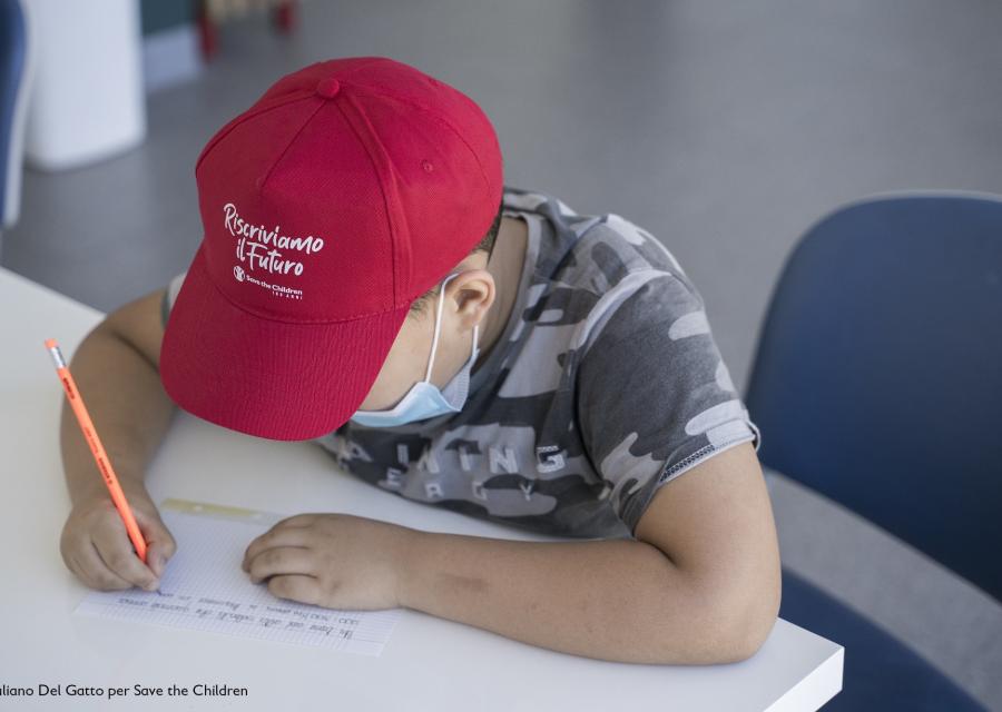 bambino seduto alla scrivania mentre scrive con cappellino rosso in testa e maglietta verde