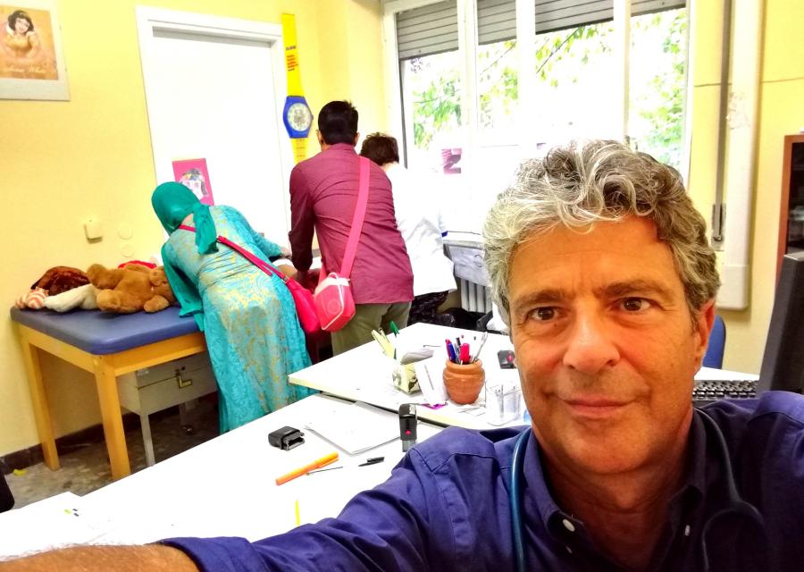 Foto selfie di un pediatra volontario nei nostri Spazi Mamme. Sullo sfondo tre mamme che cambiano un neonato nel fasciatoio.
