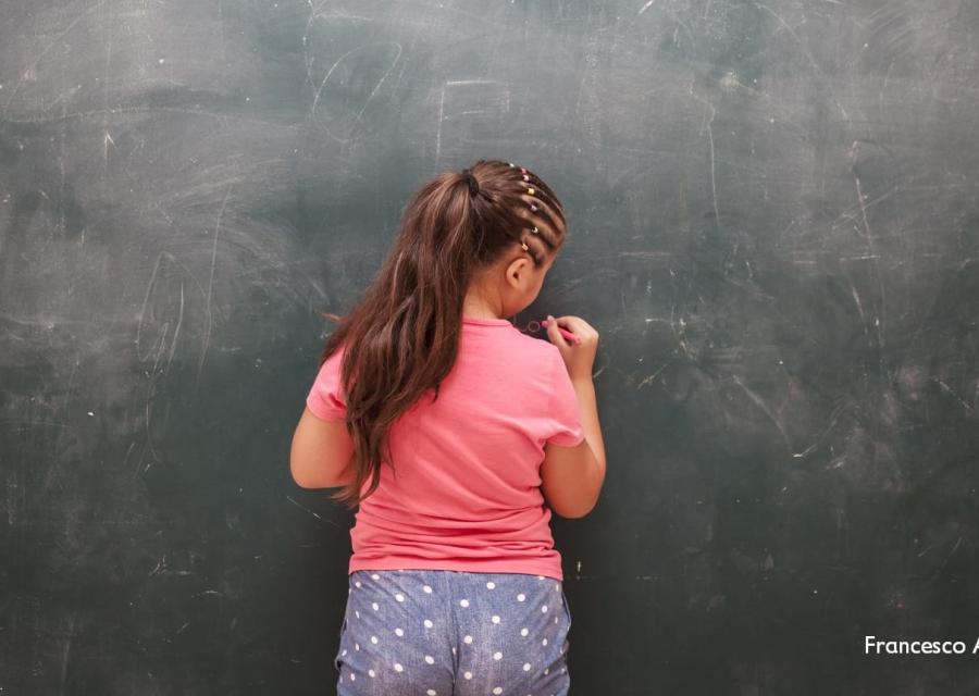 Bambina di spalle con maglietta rosa scrive alla lavagna