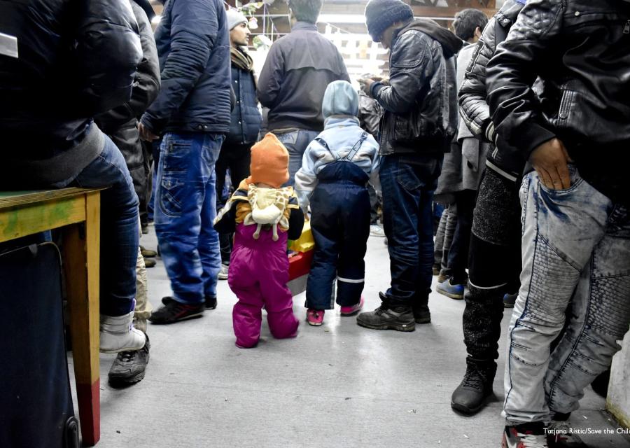 Due bambini molto piccoli di spalle sono in un ambiente chiuso molto affollato di cui si vedono soltanto le gambe e i piedi.