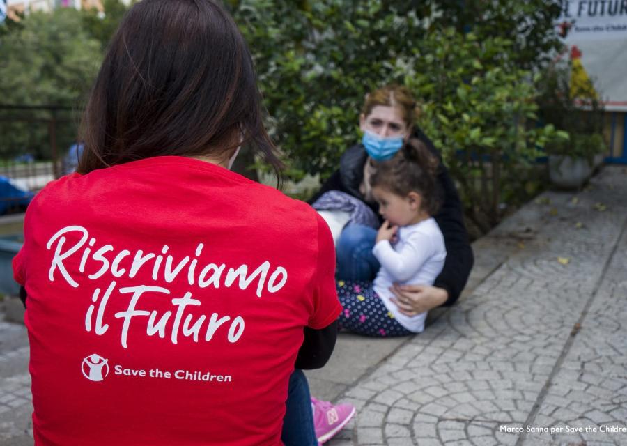 Una operatrice save the children è di spalle e indossa una maglietta save the children della campagna riscriviamo il futuro. Si trova in un cortile e davanti a lei ci sono una mamma con la sua bambina abbracciate.