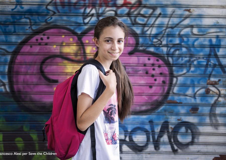 bambina con maglietta bianca e zaino viola sorride davanti a un murales