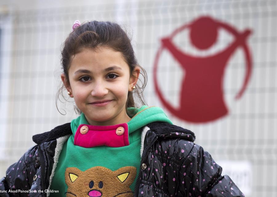 bambina sorridente con maglioncino verde e giacca, sullo sfondo il logo di save the children