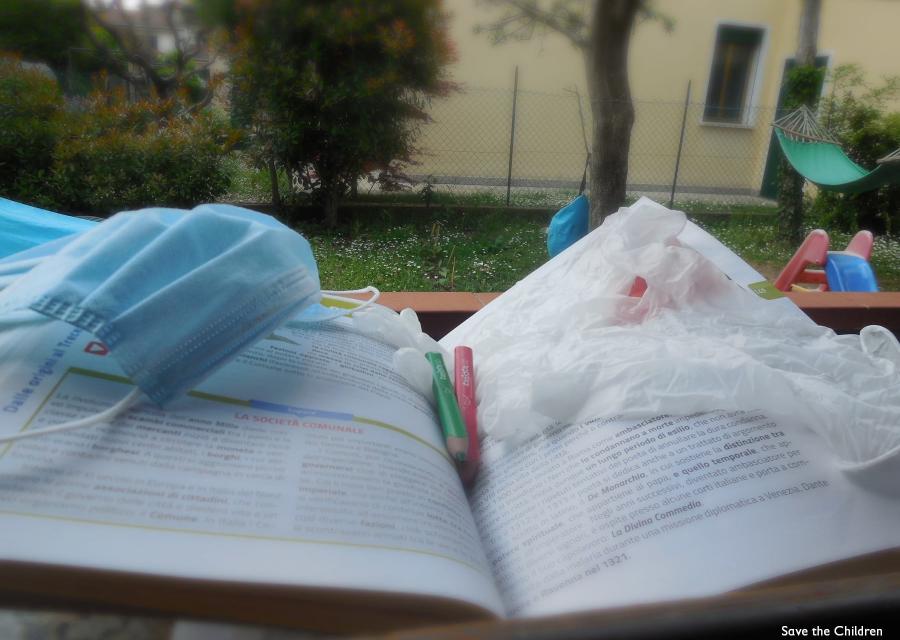 Mascherina, guanti e matite sopra un libro scolastico aperto. Sullo sfondo un giardino