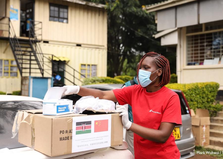 operatrice keniana di save the children con treccine e capelli rossi indossa la mascherina e sta in piedi di profilo per prendere alcuni oggetti da una scatola poggiata su un piano davanti a lei. La ragazza indossa la maglietta rossa di Save the Children