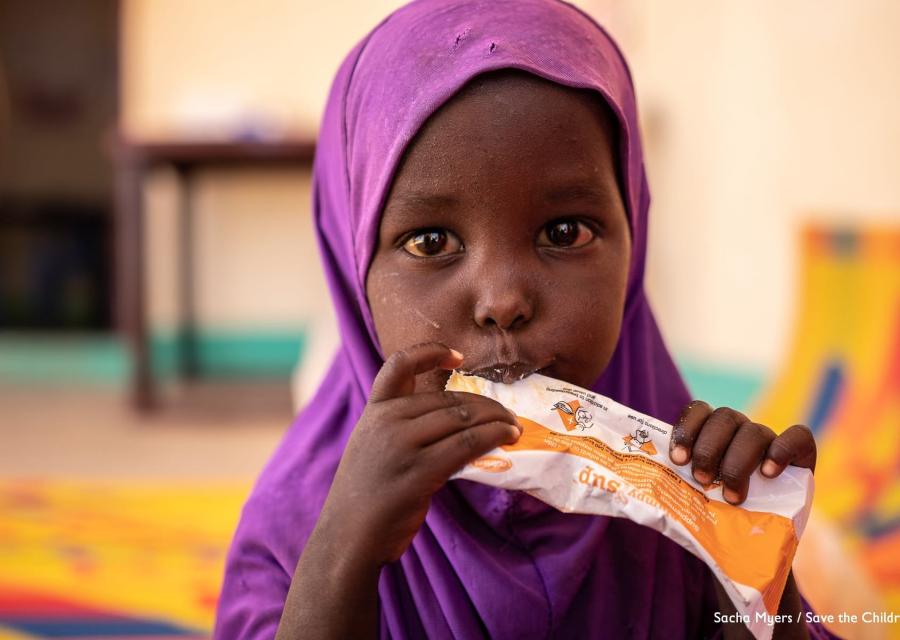 Bambina somala di pochi anni con chador viola in testa mangia una con le mani da una bustina di plumpynat