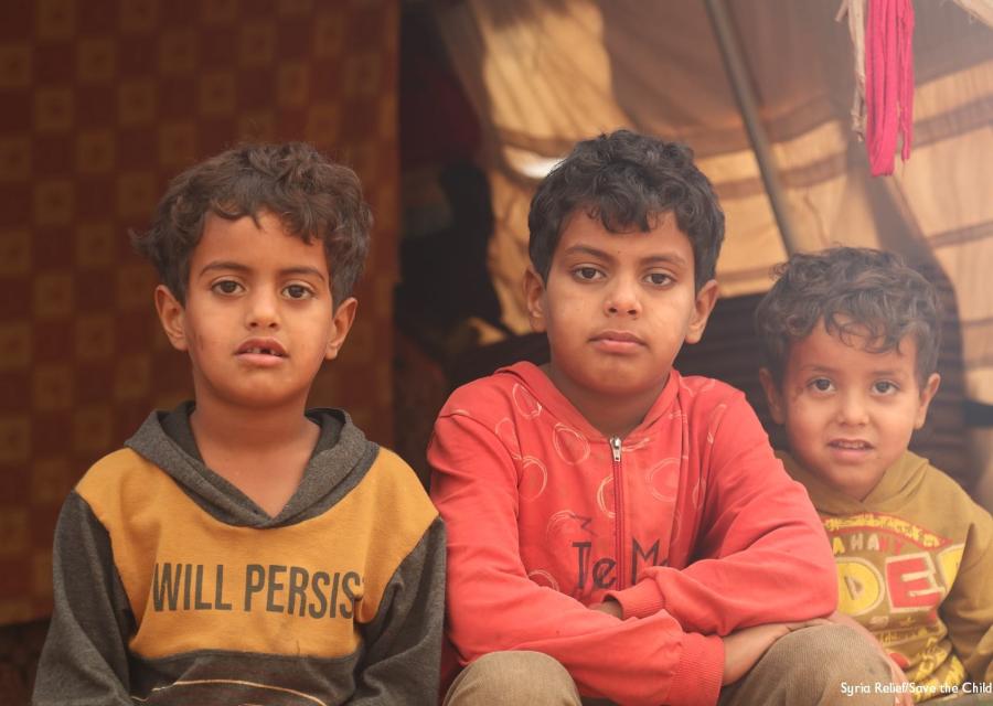 Tre bambini siriani seduti davanti alla tenda del campo in cui vivono guardano in camera. Il taglio è di mezzo busto