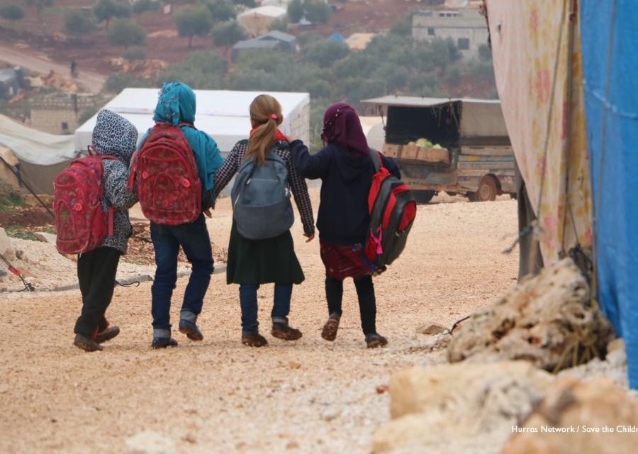 quattro bambini siriani camminano di spalle con gli zaini sulle spalle in un campo per sfollati