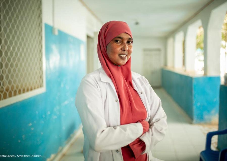 Operatrice sanitaria Fadumo che sorride con camice bianco e vero rosa attorno al volto con braccia incrociate mentre lavora nel nostro centro per la cura dei bambini malnutriti in Somalia