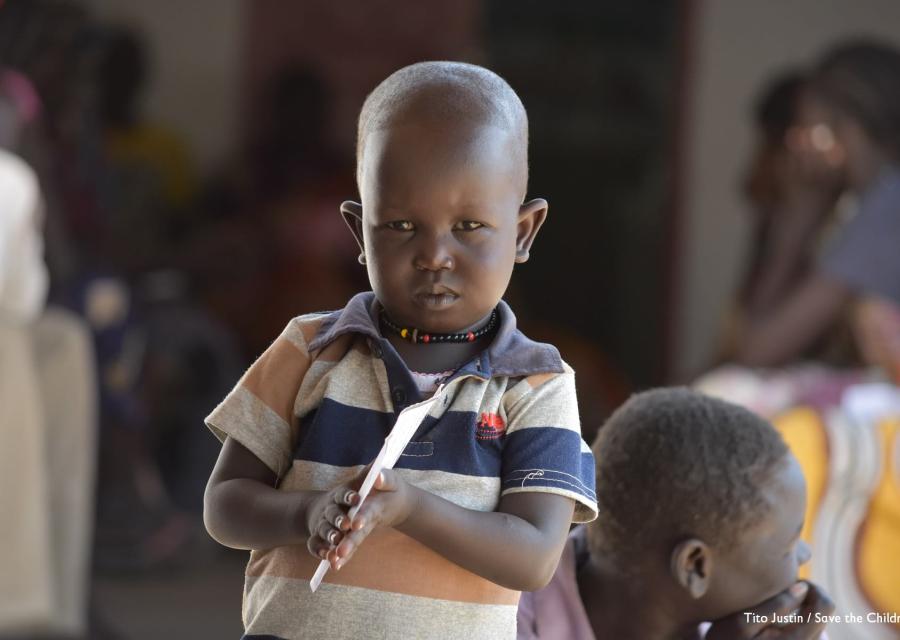 Mezzo busto di un bambino sudanese con maglietta a righe arancioni blu e bianche che tiene con due mani un foglio