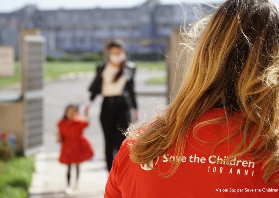oratrice save the children di spalle e davanti a lei sfocate una donna tiene per mano una bambina