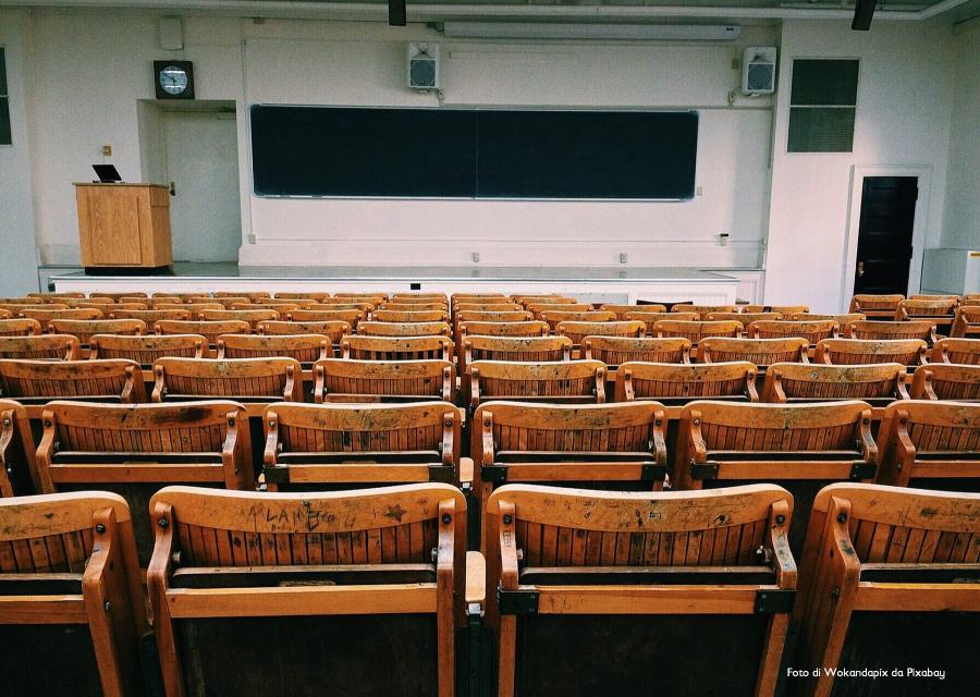 Aula universitaria vuota con sedie di legno pieghevoli, lavagna e leggio su un rialzo.