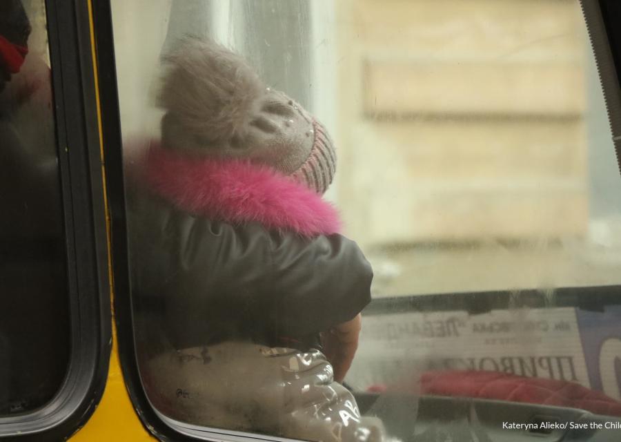 una bambina di spalle seduta su un autobus si vede dal finestrino. La bambina è vestita con un giubbotto pesante e un cappello di lana.
