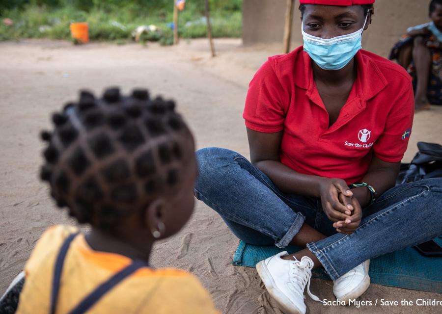 un operatrice save the children in Mozambico è seduta a terra a gambe incrociate e guarda una bambina di spalle seduta di fronte a lei.