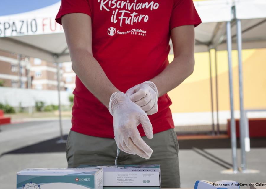 Primo piano del mezzo busto di un operatore Save the Childrenche indossa una maglietta rossa a maniche corte con la scritta del nome del progetto sostenuto: Riscriviamo il futuro. L uomo indossa inoltre un paio di guanti monouso ed è ripreso nel gesto di sfilarli e appoggiarli su un tavolino davanti a lui. Il volto non è inquadrato.