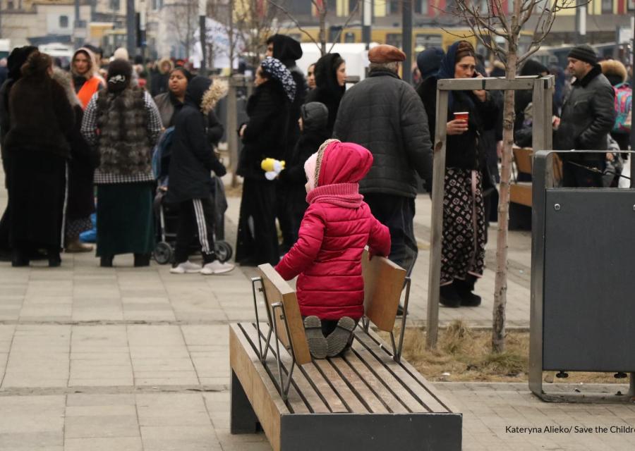 Una bambina con cappotto rosa di spalle è in ginocchio su una panchina. Davanti a lei una folla di persone.