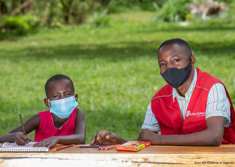 Un bambino ugandese è seduto alla scrivania all aperto e accanto lui è seduto un operatore save the children.