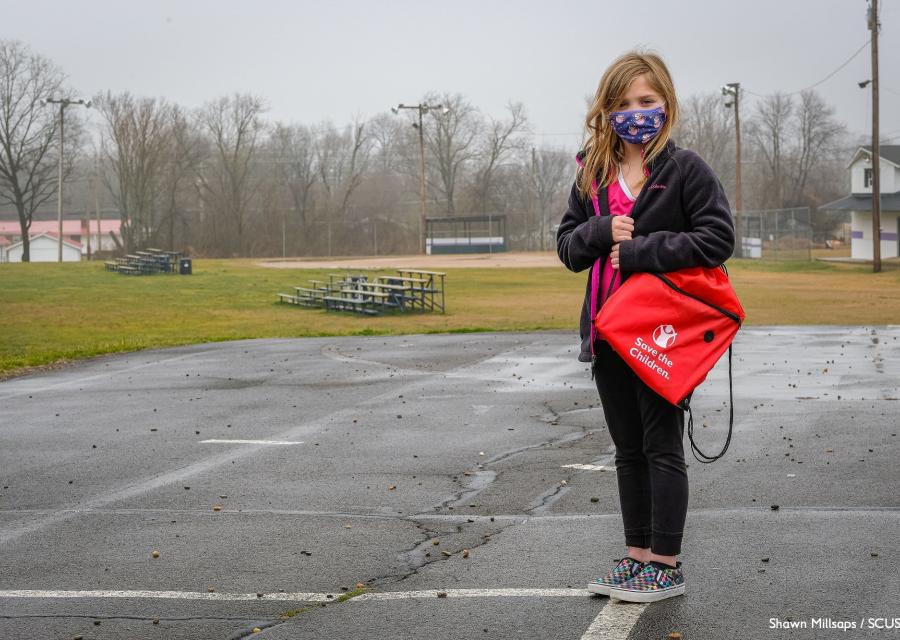 bambina in piedi in un parcheggio con mascherina per proteggersi dal Covid-19 tiene in spalla uno zaino scolastico rosso di Save the Children