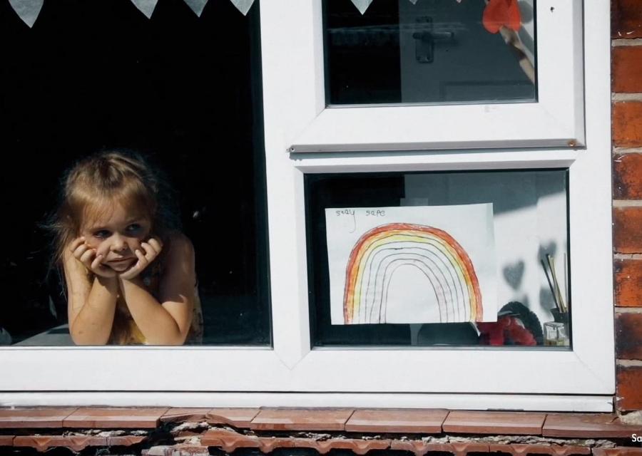 una bambina affacciata alla finestra guarda in basso pensosa. Sulla destra è appeso alla finestra il disegno di un arcobaleno
