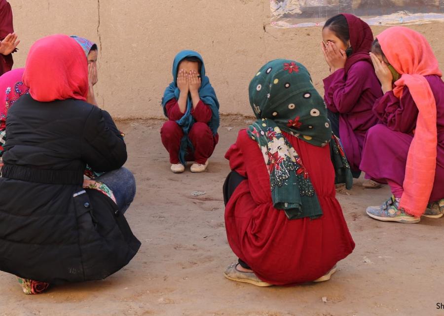 bambine vestite in viole e rosso sono sedute in cerchio e si coprono il volto con le mani.