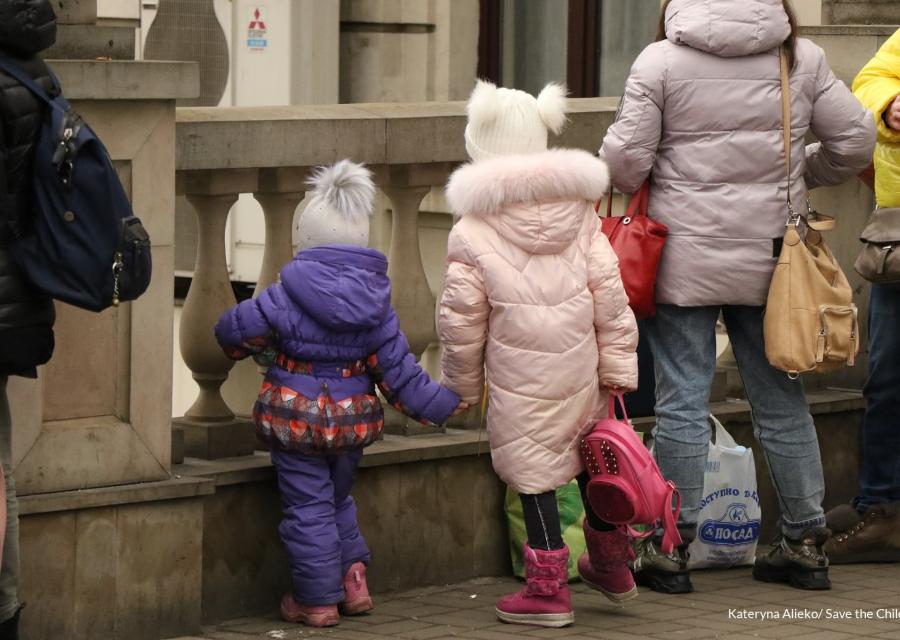 Due bambine di spalle con i giacconi si danno la mano e guardano avanti affacciate a una balaustra.
