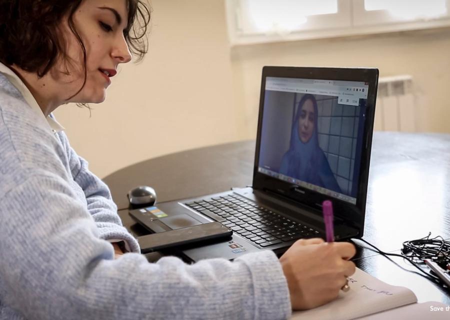 Una ragazza seduta alla scrivania di profilo scrive su un quaderno e davanti a un pc nel cui schermo c è una video chiamata