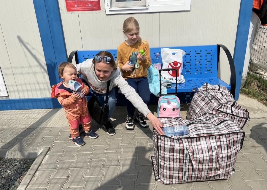 mamma con due bimbi su una panchina blu con logo save the children italia con una busta con aiuti umanitari