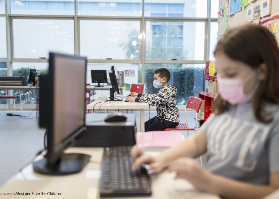 bambina in primo piano e bambino in secondo piano entrambi seduti alla scrivania mentre scrivono al PC