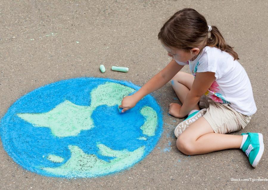 Una bambina bionda con la coda è seduta a terra sulla strada e disegna con i gessi un mappamondo. Lei è vestita con una maglietta a maniche corte bianca e dei pantaloncini marroni.