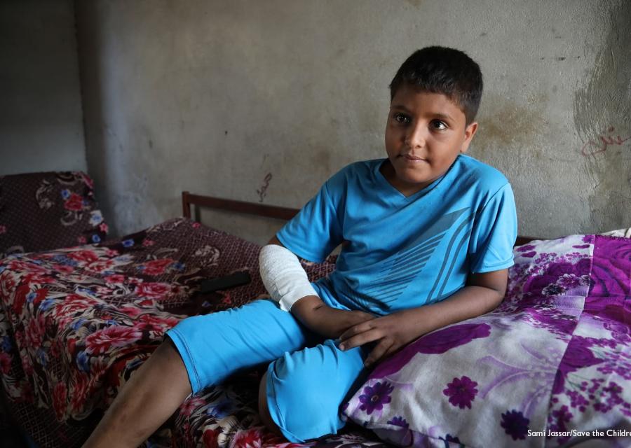 Un bambino yemenita seduto sul letto indossa una tuta celeste a maniche corte e pantaloncini. Ha una fasciatura al braccio e alla gamba
