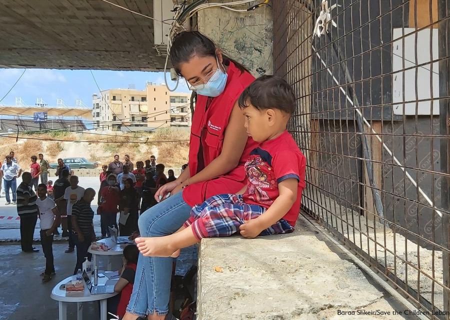 operatrice save the children con mascherina seduta su un muretto a fianco di un bimbo. Sullo sfondo varie presone che camminano. 