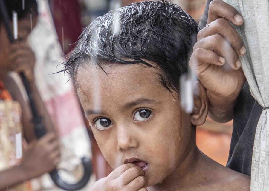 Un bambino rohingya si protegge dalla pioggia sotto un telo tenendo per mano la madre.