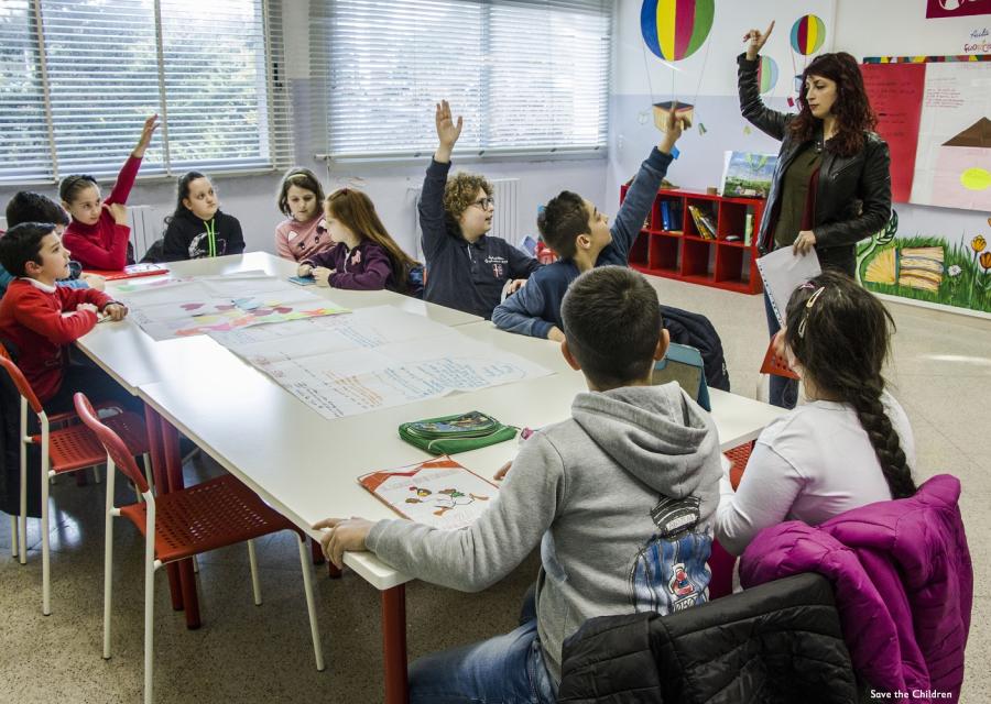 una decina di bambini seduti a un banco di scuola alzano la mano alla domanda della maestra