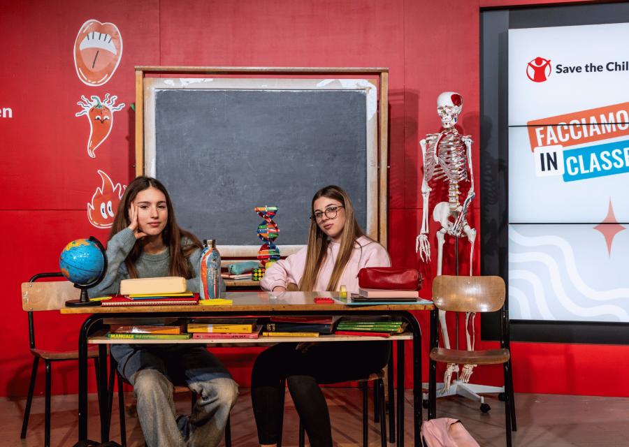 Due ragazze sedute dietro banco e sullo sfondo lavagna