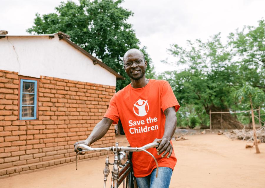 ragazzo del Malawi di fronte una casa con maglietta rossa Save the Children e tra le mani il manubrio di una bici 