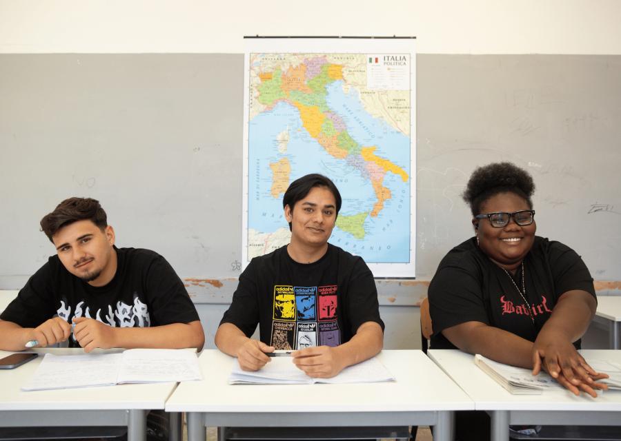 Studentessa e studenti con background migratorio sorridenti su banco di scuola con dietro la cartina d'Italia