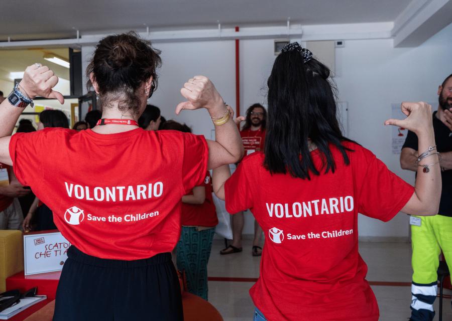 Due volontarie di Save the Children di schiena che indicano la scritta sul retro della maglietta "volontario" con logo Save the Children