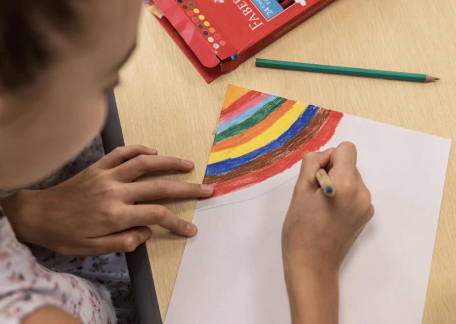 bambina dall'alto che tiene in mano una matita colorata e disegna un arcobaleno su un banco di scuola