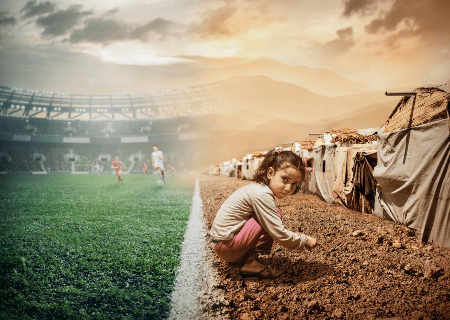 A sinistra bambina rannicchiata a bordo campo di calcio a destra calciatori sullo sfondo 