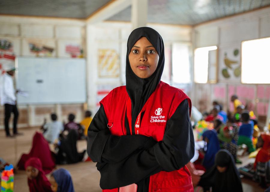 Primo piano di una operatrice con pettorina rossa di Save the Children all'interno di uno Spazio a Misura di Bambino in Somalia. Sullo sfondo, bambini all'interno dello spazio svolgono un'attività guidata ad un altro operatore