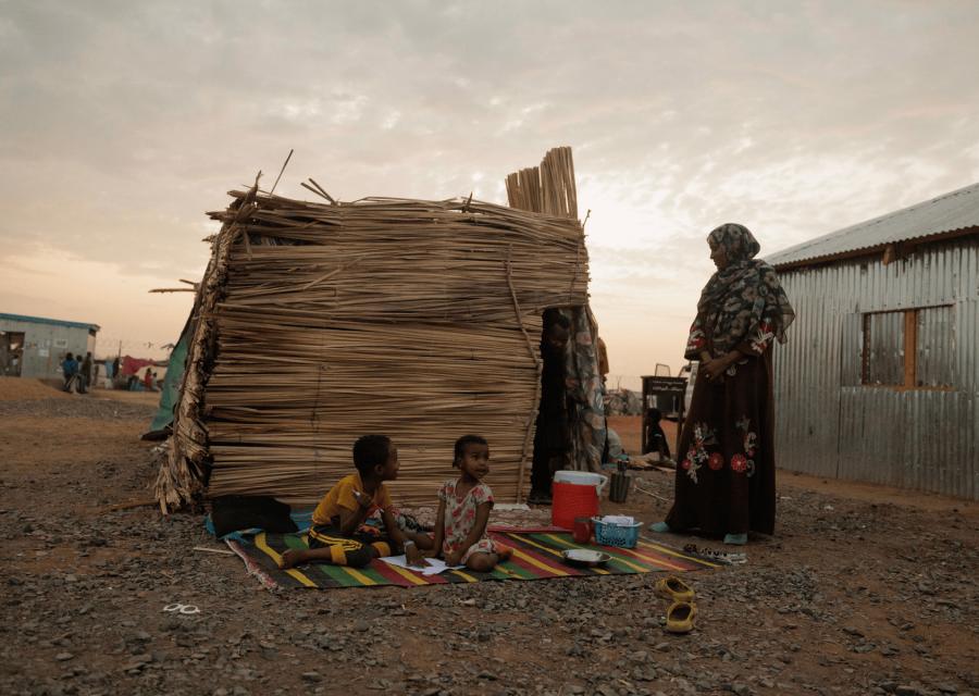 Famiglia in Sud Sudan con bambini seduti su una stuoia a terra e madre in piedi accanto a loro con sullo sfondo la loro casa