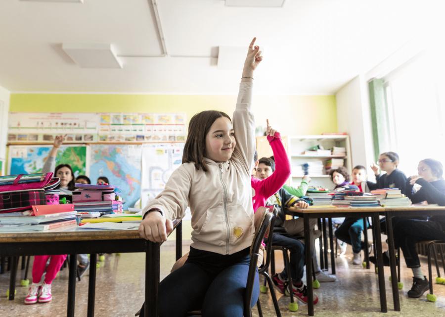 Bambina sui banchi di scuola che alza la mano con dietro altri bambini a scuola