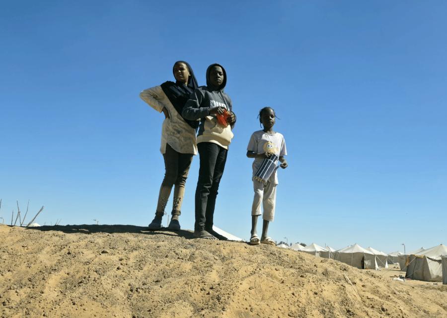 Tre minori Sudanesi in piedi sulla sabbia davanti a un campo per rifugiati.
