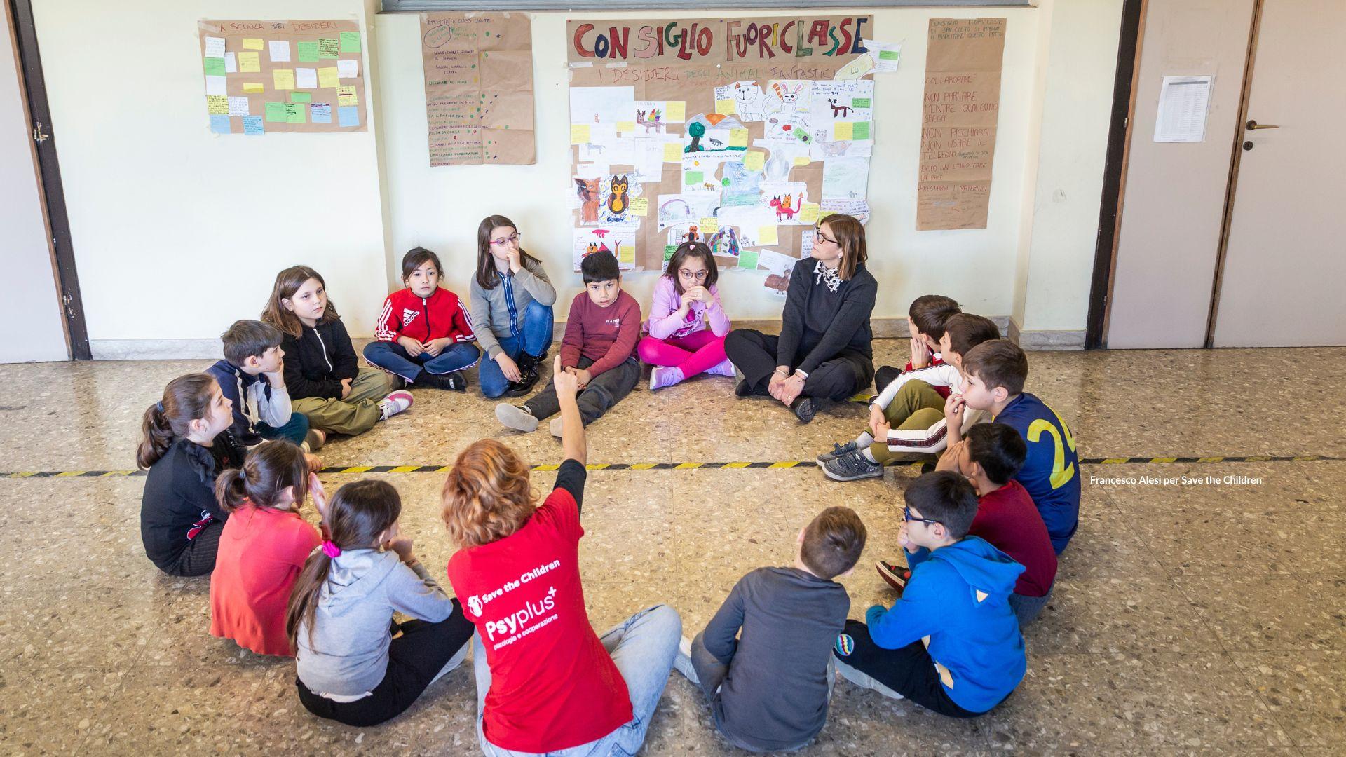 Bambini seduti per terra, in cerchio, durante il consiglio Fuoriclasse, con operatrice Save The Children e insegnante.