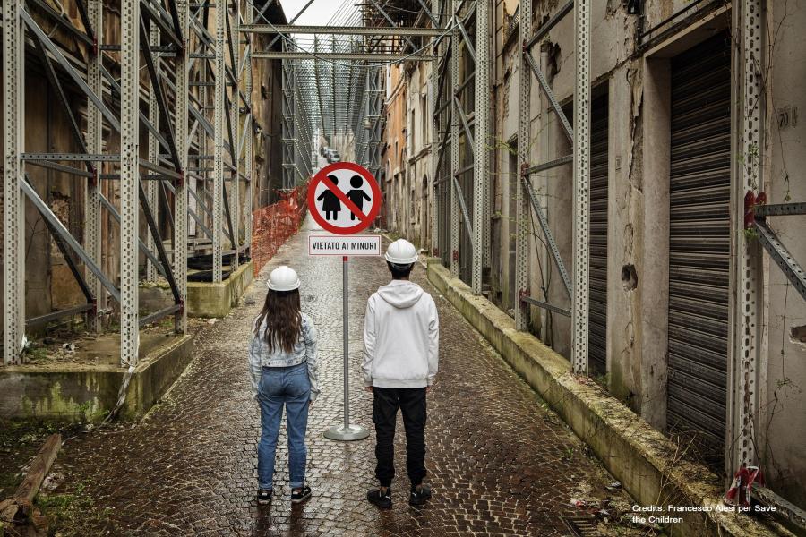 2 bambini nella zona rossa a L'Aquila con caschetto bianco di spalle davanti a cartello vietato ai minori