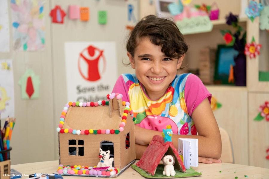 bambina seduta a un banco di scuola con di fronte la sua creazione di cartone guarda verso di noi sorridente
