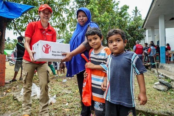 un operatore di Save the Children consegna a una mamma e a due bambini un pacco contenente un kit igienico