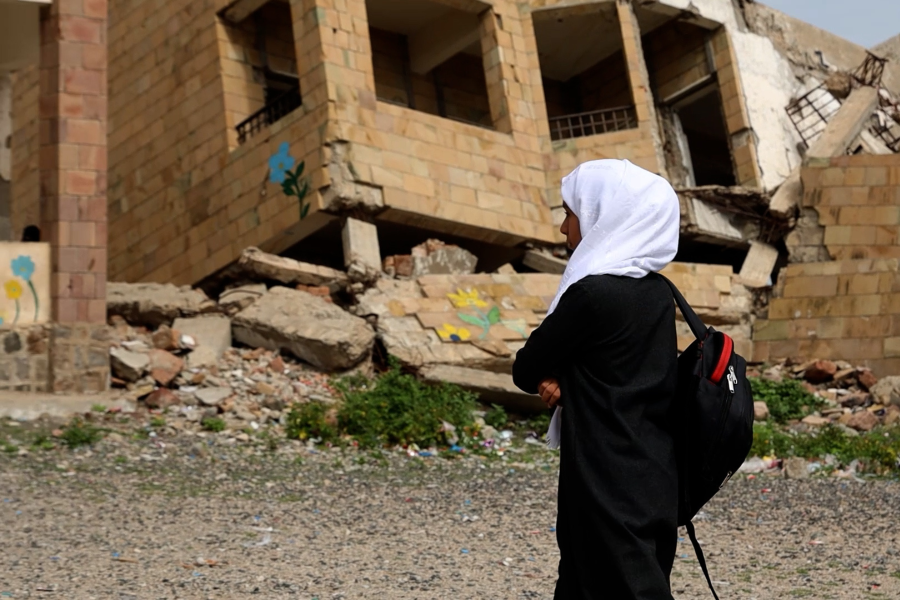 Bambina in Yemen con zaino sulle spalle di fronte a un palazzo bombardato